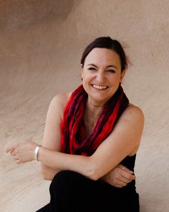 Andrea Bacher-Steiner lacht in die Kamera, mit rotem Schal vor hellem Sandstein