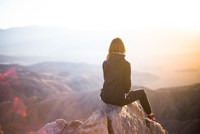 Frau sitzt auf einem Felsen und blickt über eine Berglandschaft im Gegenlicht