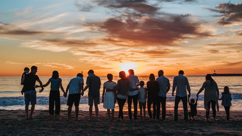 Eine Gruppe von Menschen steht als Silhouette im Abendlicht am Meer, teils Hand in Hand