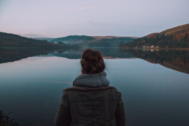 Person mit Schal steht von hinten gesehen am Ufer eines ruhigen Sees, dahinter bewaldete Hügel